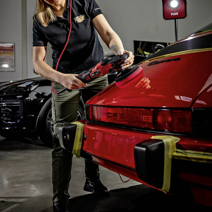 A woman in a black polo shirt and olive pants uses a red orbital sander to apply clear coat to the rear trunk of a red sports car in a garage setting.