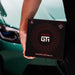 A person holding a black GT1 Easy-Apply Ceramic Coating Kit box in front of a teal car.