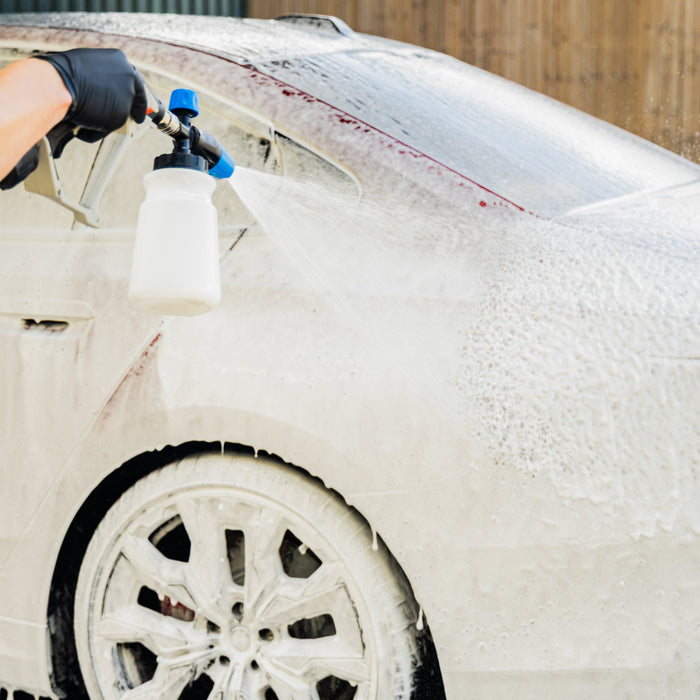 A close-up of a red car covered in snow foam being sprayed with a foam cannon by a person wearing a black glove, with a wooden fence and building in the background.