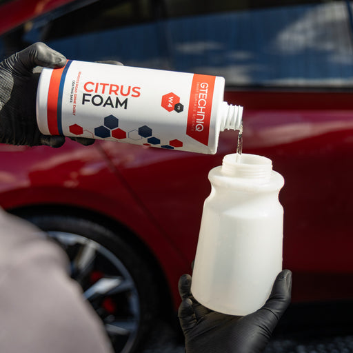 A person wearing black gloves pours liquid from a white bottle labeled 'CITRUS FOAM' and 'GTECHNIQ' into a white container, with a red car blurred in the background.