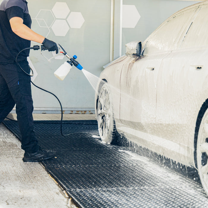 A man in a black uniform sprays a red car with a foam cannon at an outdoor car wash bay.