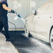 A man in a black uniform sprays a red car with a foam cannon at an outdoor car wash bay.